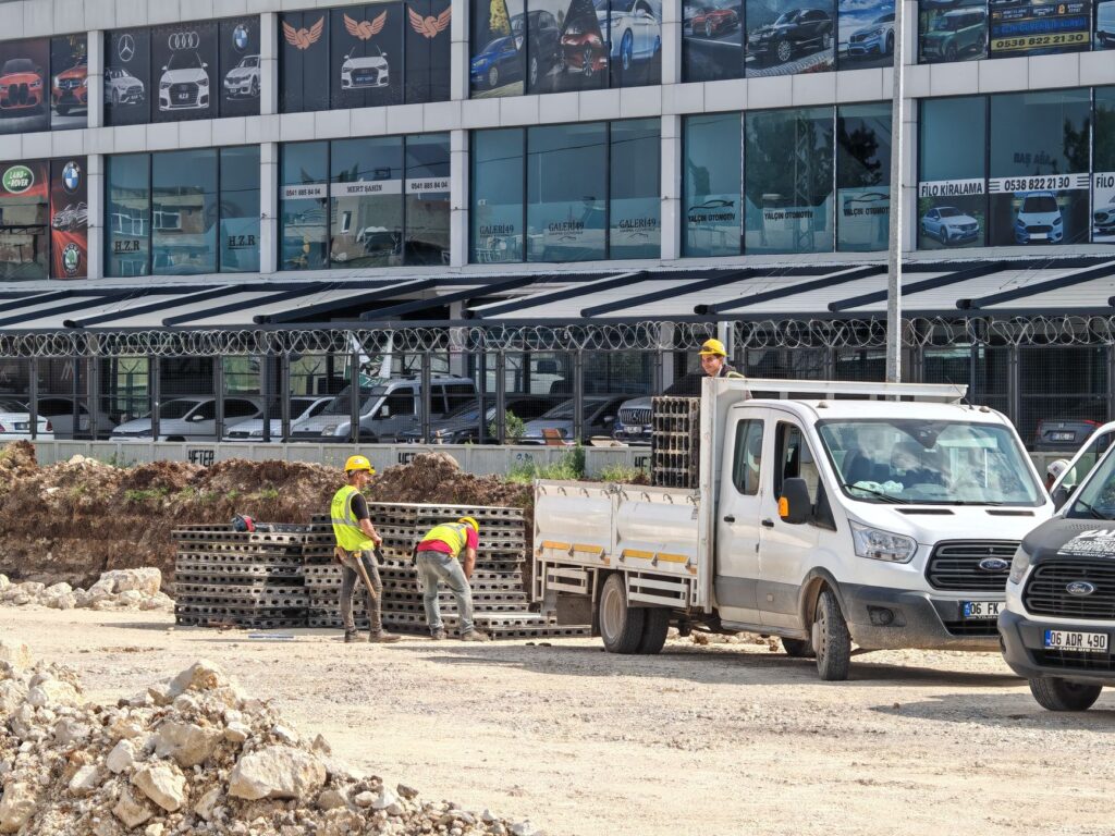 Workers unloading materials from a truck at a construction site in front of a car dealership.