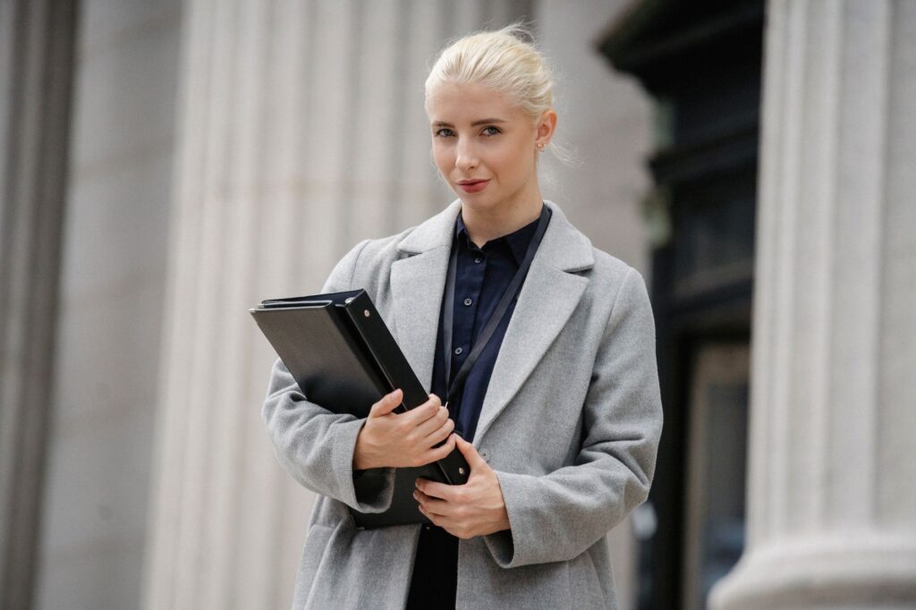 Positive young businesswoman wearing stylish formal outfit standing with documents in hands outside stone building and looking at camera