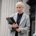 Positive young businesswoman wearing stylish formal outfit standing with documents in hands outside stone building and looking at camera