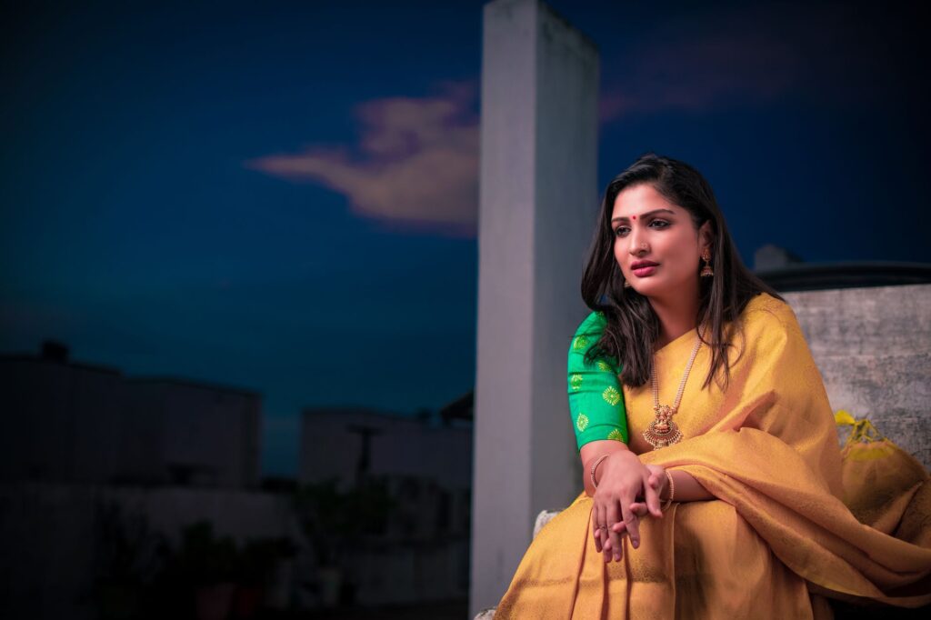 Portrait of an Indian woman in a yellow sari, seated with a serene expression outdoors during twilight.