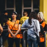 A lively scene with soccer fans and stewards interacting outside a stadium on a sunny day.