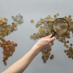 Top view of crop unrecognizable traveler with magnifying glass standing over world map made of various coins on gray background