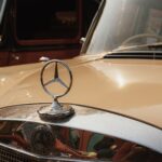 Close-up of a vintage Mercedes-Benz hood ornament on a classic car with reflections.
