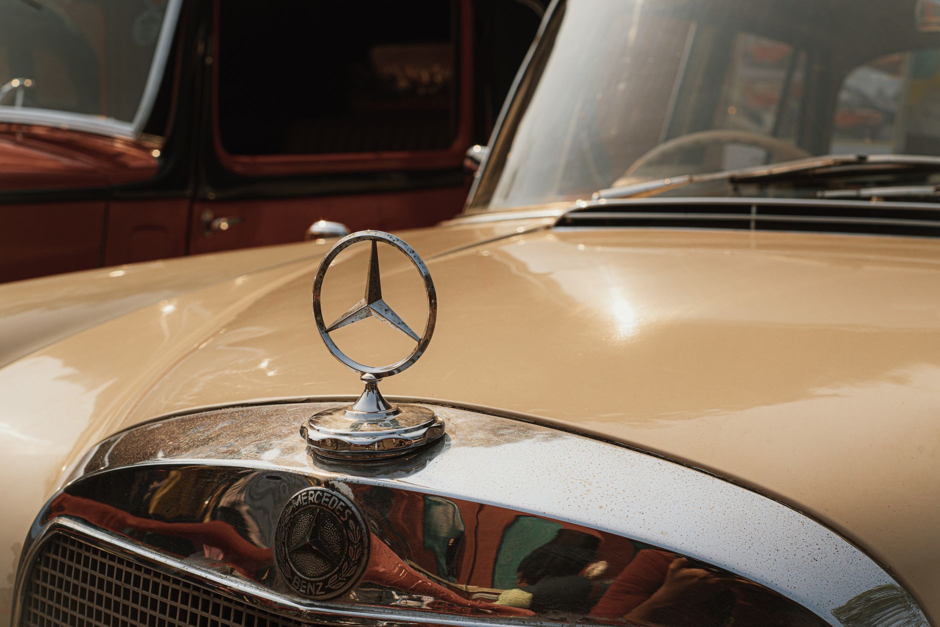Close-up of a vintage Mercedes-Benz hood ornament on a classic car with reflections.