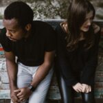 A couple sitting apart on a park bench, expressing emotions. Outdoors setting.