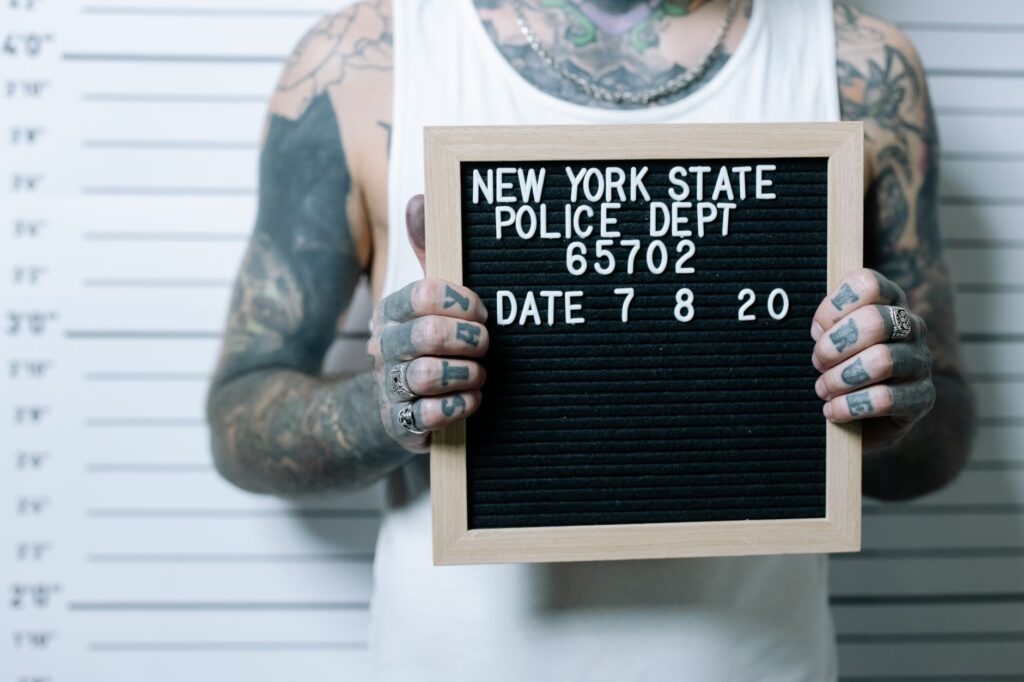 Close-up of tattooed man holding a mugshot sign in front of a height chart.