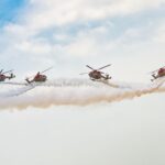 Four helicopters perform synchronized aerobatics with smoke trails in the sky.