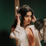 A woman wearing headphones recording vocals in a music studio with a microphone.