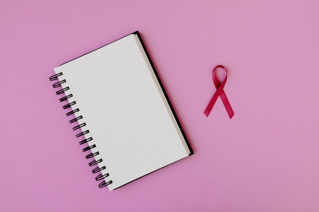 Open spiral notebook with pink breast cancer awareness ribbon on a pink background, offering copy space.