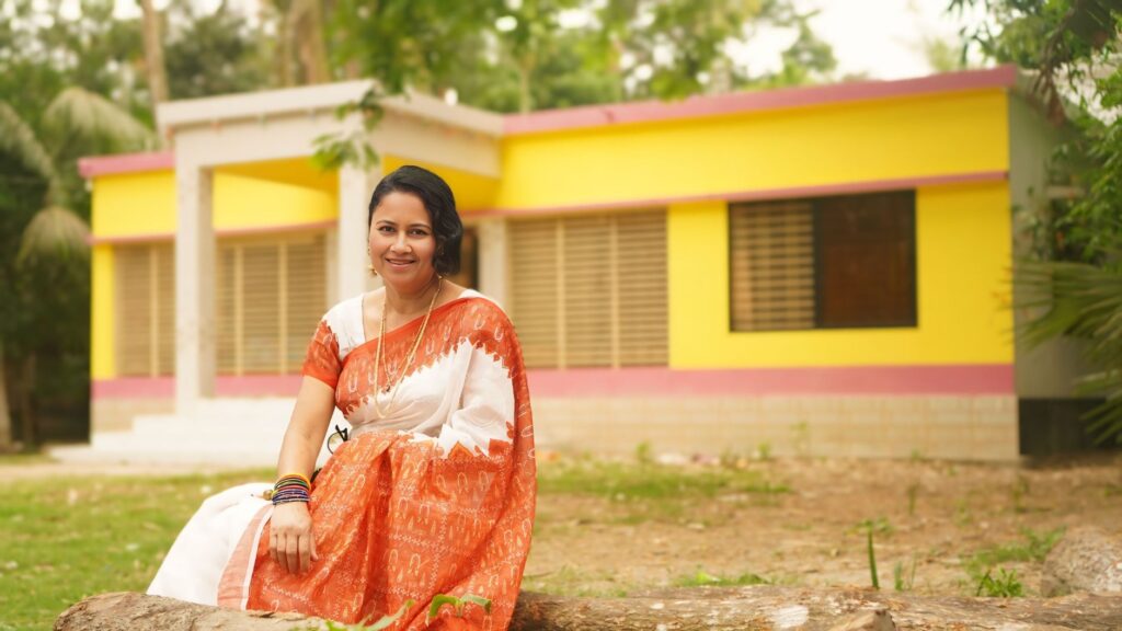 A smiling woman in a saree sits outdoors in front of a vibrant yellow house.