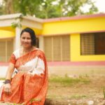A smiling woman in a saree sits outdoors in front of a vibrant yellow house.