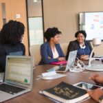 Four diverse women engaged in a business meeting with laptops and presentations in an office.