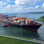 Large container ship maneuvered by tugboats through the Panama Canal under a clear sky.