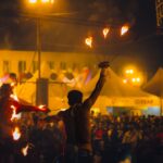 Crowd mesmerized by fire dancers under vibrant festival lights at night.