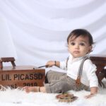 Adorable baby dressed as an artist playing with a paintbrush and easel in a cozy setting.