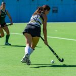 Two female athletes competing in a dynamic field hockey match on a sunny day.
