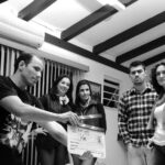 A group of adults collaborate on a filmmaking project, seen holding a clapperboard indoors.
