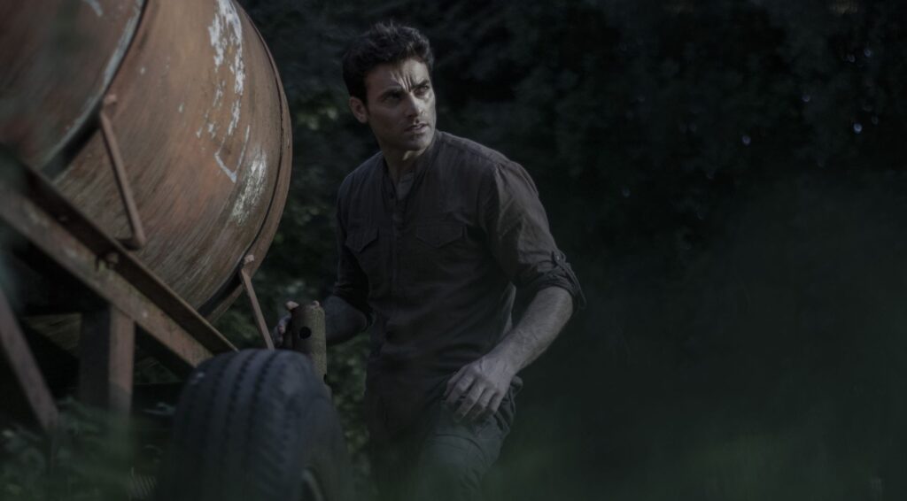 A man with short hair stands beside a rustic trailer in a forest setting.