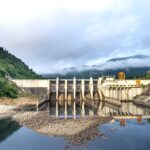 Industrial hydroelectric power plant located near river and stony coast against green mountain range covered with mist on summer day