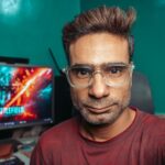 A man with glasses poses for a selfie in his home office, with a computer screen in the background.
