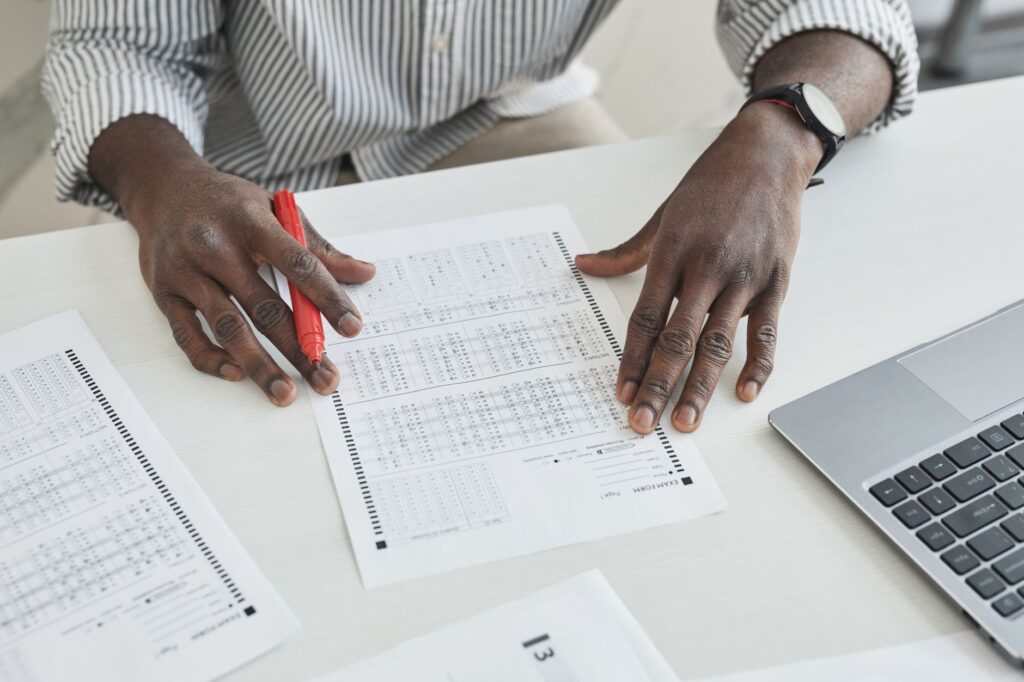 An educator carefully checks exam papers using a red marker at a desk, illustrating focused grading.