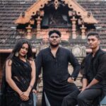 Three adults in traditional Indian attire pose elegantly in front of a cultural building.