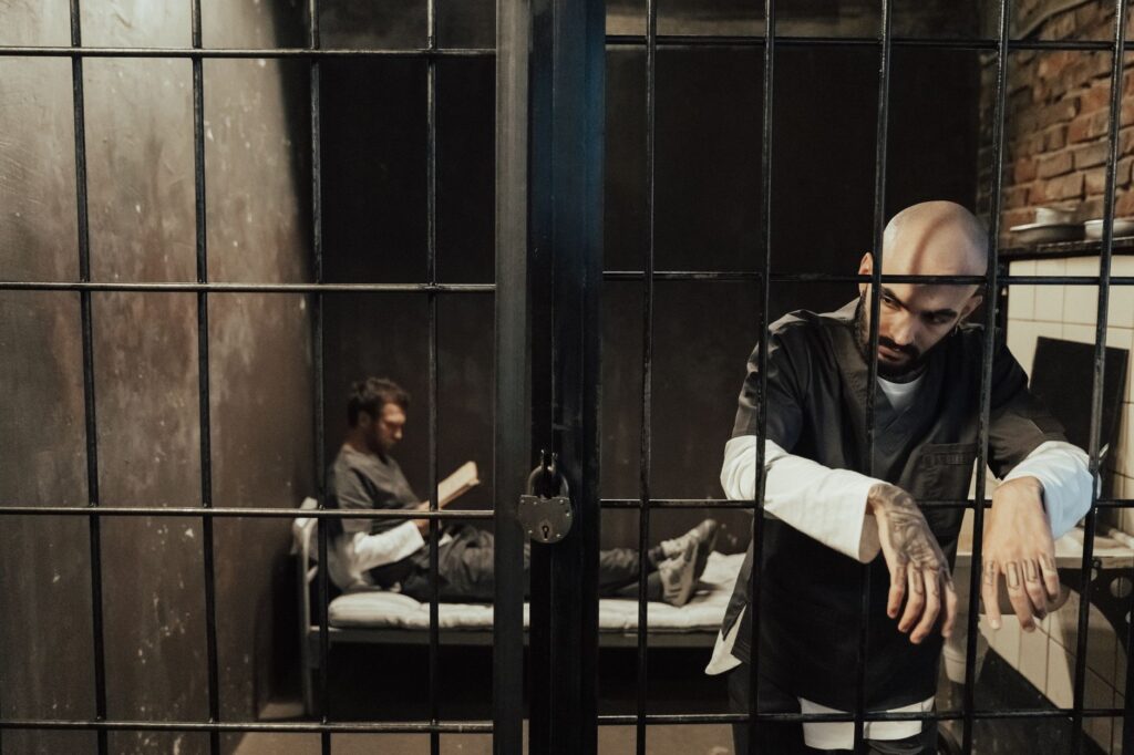 Two men in a dimly lit prison cell, one reading on bed, the other standing somberly by bars.