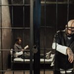 Two men in a dimly lit prison cell, one reading on bed, the other standing somberly by bars.
