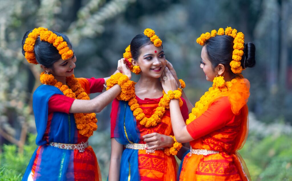 Three women in traditional Bengali attire, adorned with marigold flowers, celebrate outdoors.