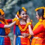 Three women in traditional Bengali attire, adorned with marigold flowers, celebrate outdoors.