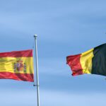 Flags of Spain and Belgium waving on poles under a blue sky, symbolizing international relations.