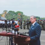 Side view of mature colonel with order standing near microphones on asphalt pavement behind army of soldiers near airplane monument under serene sky during national military holiday and looking away