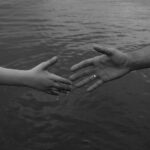 Monochrome image of two hands reaching out over water, symbolizing connection and support.