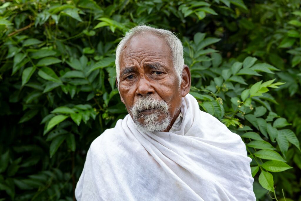Portrait of an elderly man wearing traditional clothing in a rural Indian setting with lush greenery.