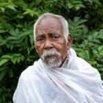 Portrait of an elderly man wearing traditional clothing in a rural Indian setting with lush greenery.