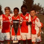 Young soccer players celebrate winning the 2023 SEJG Cup with their trophy outdoors.