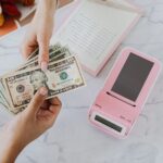 Close-up of hands exchanging US dollars with a pink calculator on a marble surface.