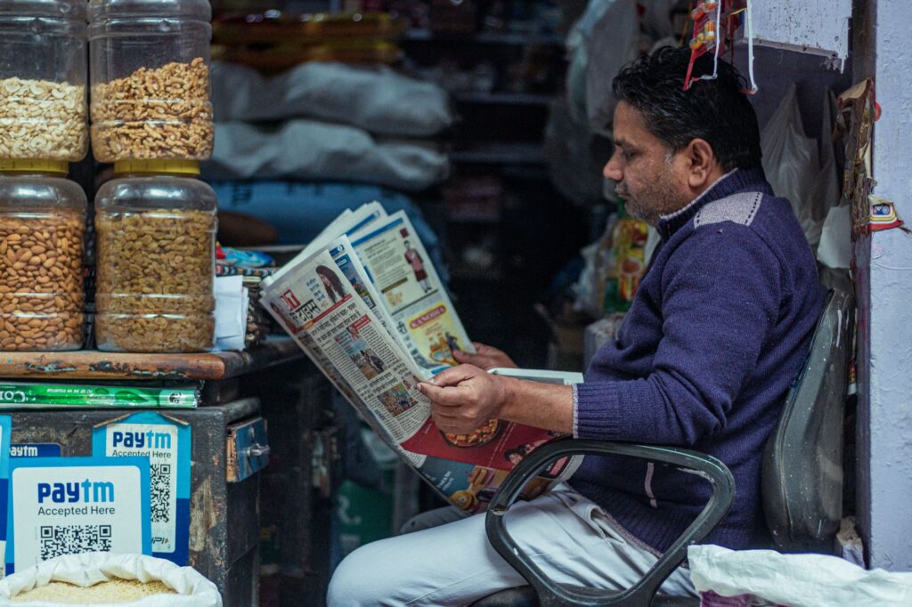 Man reading a newspaper in a small shop, surrounded by jars of snacks. Indoor market scene.