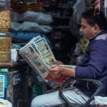 Man reading a newspaper in a small shop, surrounded by jars of snacks. Indoor market scene.