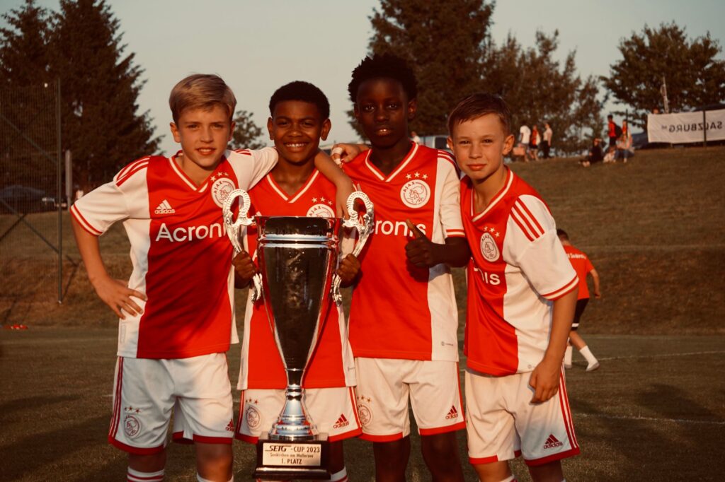 Young soccer players celebrate winning the 2023 SEJG Cup with their trophy outdoors.