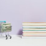 A shopping cart with dollar bills next to a stack of textbooks on a light background.