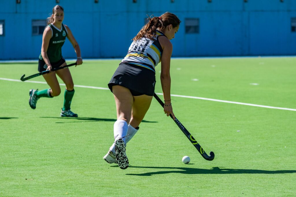 Two female athletes competing in a dynamic field hockey match on a sunny day.
