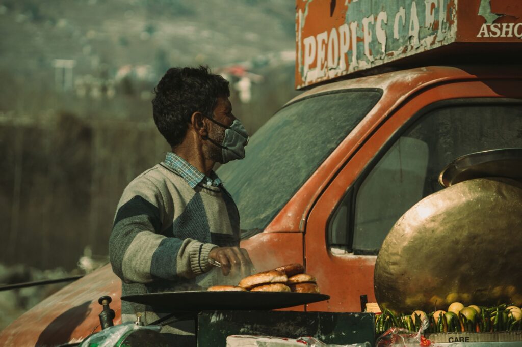 A street vendor wearing a mask prepares food outdoors, reflecting the ongoing pandemic.