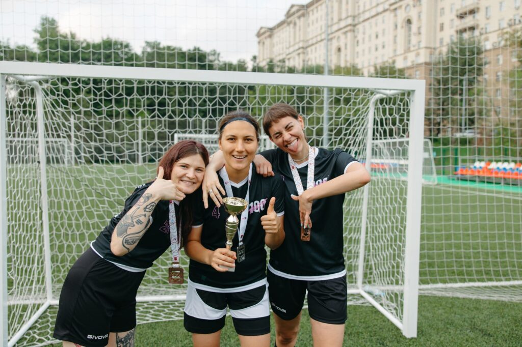 Three female soccer players celebrate their win with a trophy on an outdoor field, displaying joyful expressions and team spirit.