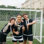 Three female soccer players celebrate their win with a trophy on an outdoor field, displaying joyful expressions and team spirit.
