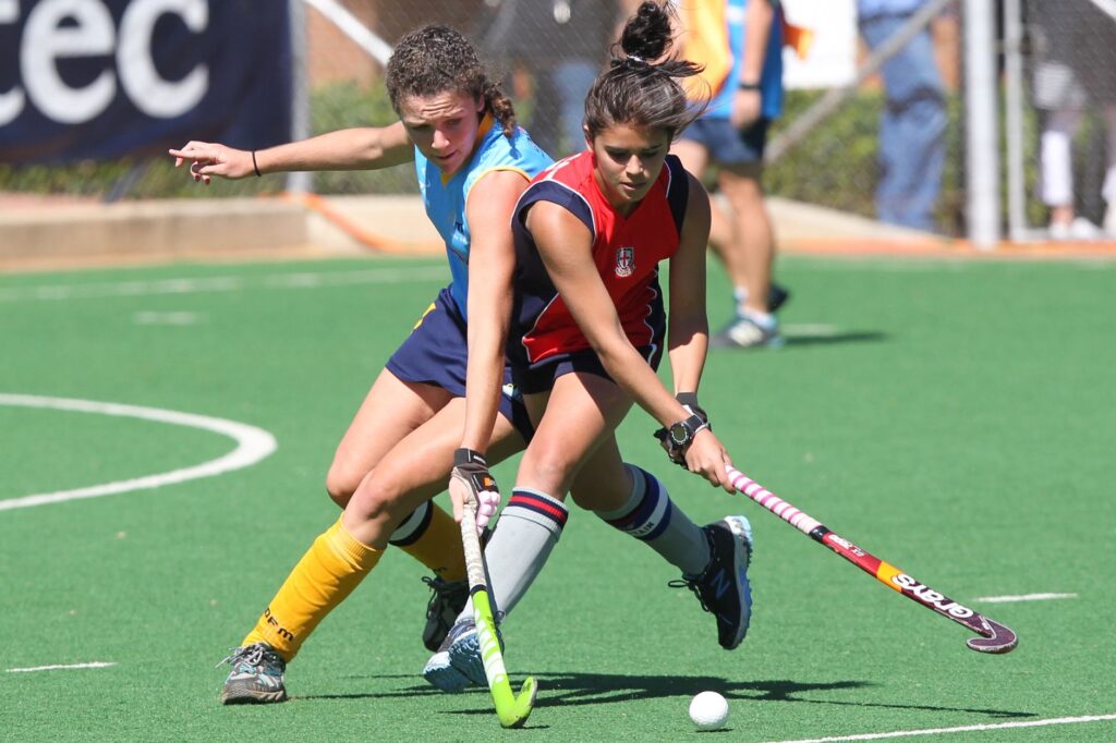 Two female field hockey players compete fiercely on an outdoor turf field.