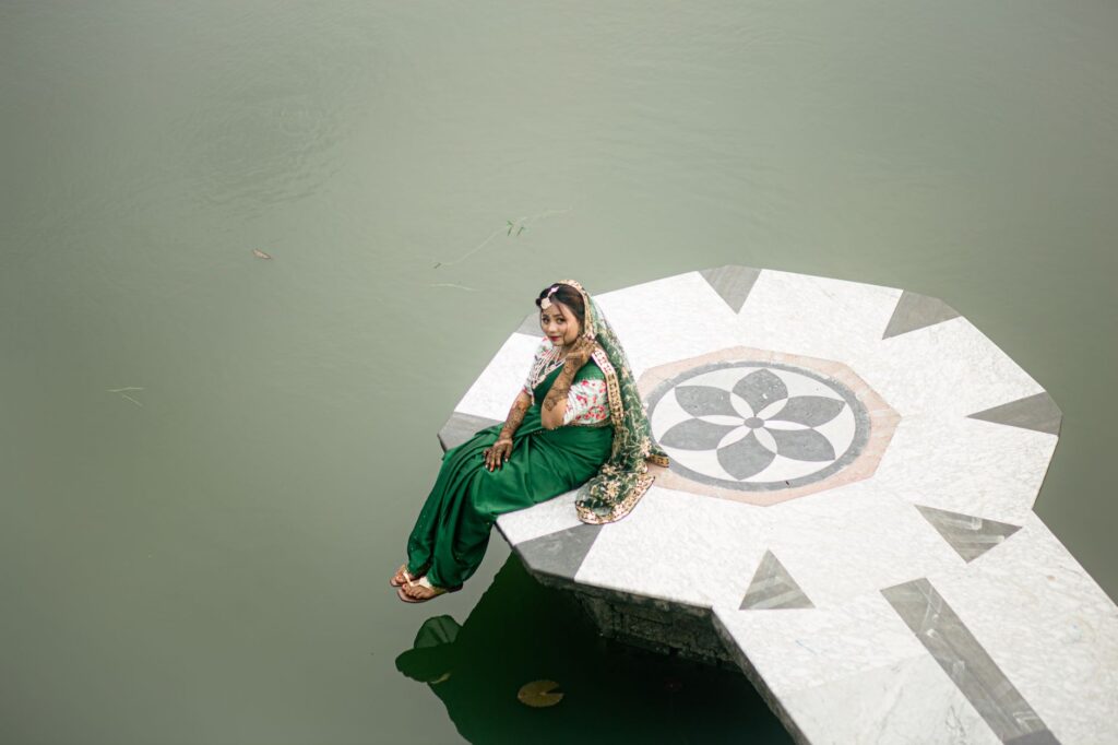 A woman in vibrant traditional Bangladeshi attire sits gracefully on a platform over water.