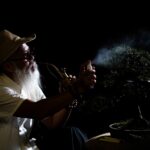 Elderly man sprays a bonsai tree in a dramatic, dark setting with low key lighting.