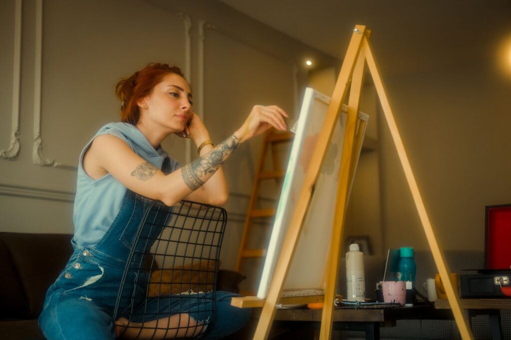 Red-haired artist sitting indoors, painting on canvas in a cozy home setting.