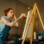 Red-haired artist sitting indoors, painting on canvas in a cozy home setting.